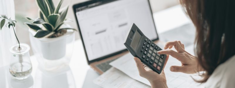 A person at a desk with a laptop and accounting documents in front of them doing calculations on a calculator.