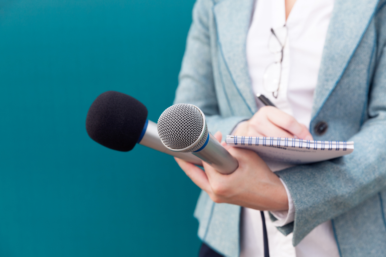 A journalist holding two microphones while writing notes down in a notepad.